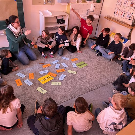 Ein Klassenzimmer mit Kindern auf dem Teppich sitzend. Eine Frau leitet den Unterricht. Karten mit Wörtern sind auf dem Teppich verstreut. Die Kinder sind in die Aktivität vertieft.