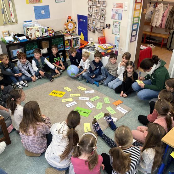 Kinder sitzen in einem Kreis um eine Weltkugel, während eine Lehrerin einen Stift hält und auf Papier schreibt. Der Raum hat Regale mit Büchern, eine Weltkugel und ein Banner an der Wand.