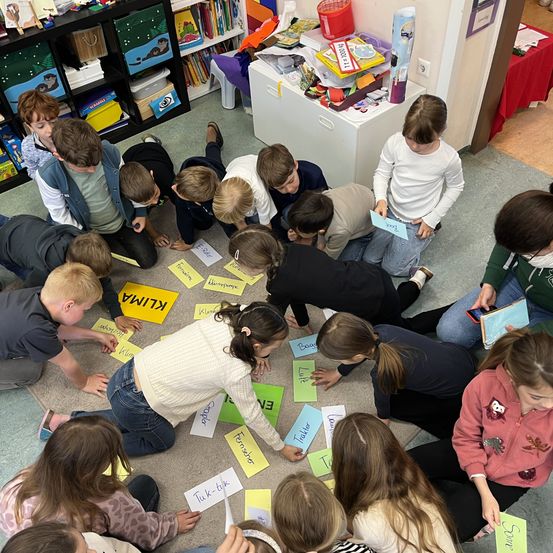 Eine Gruppe von Kindern sitzt auf dem Boden und macht wahrscheinlich eine Aktivität. Der Boden ist mit gelben Papieren bedeckt, auf denen Wörter stehen. Eine Frau in einem weißen Pullover hält ein Papier. Hinter ihnen steht ein Tisch mit Büchern und anderen Sachen.