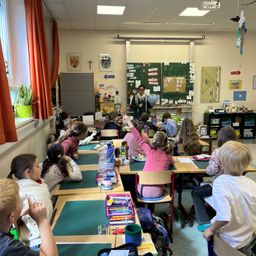 Ein Klassenzimmer mit Schülern an Tischen. Ein Lehrer steht mit einer Erdkugel vor der Klasse und interagiert mit den Schülern. Verschiedene Poster und Dekorationen schmücken die Wände.