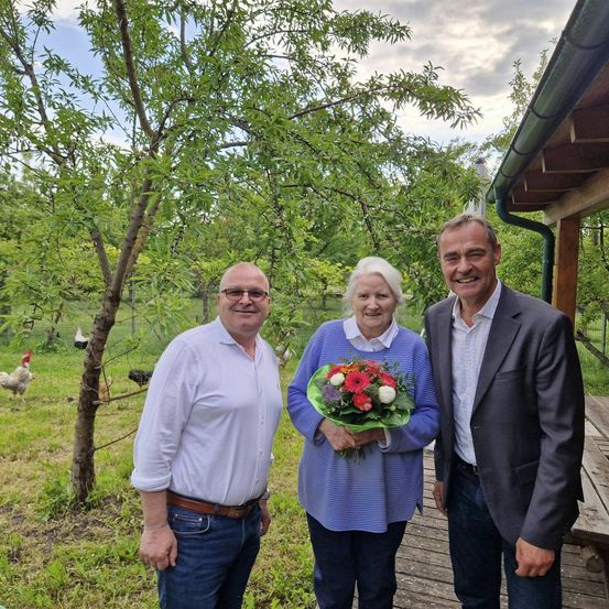 Drei Personen stehen im Freien und lächeln. Die Frau hält Blumen. Hinter ihnen steht eine Holzstruktur und Bäume. Hühner laufen in der Ferne.