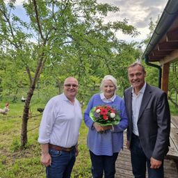 Drei Personen stehen im Freien und lächeln. Die Frau hält Blumen. Hinter ihnen steht eine Holzstruktur und Bäume. Hühner laufen in der Ferne.