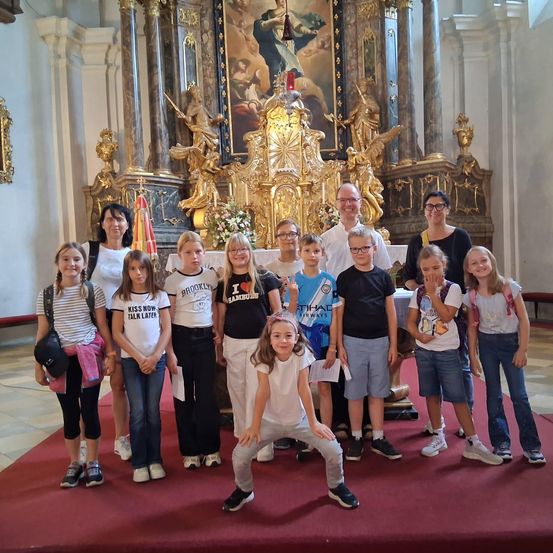 Eine Gruppe von Kindern und Erwachsenen posiert für ein Foto vor einem kunstvollen Kirchenaltar. Die Kinder tragen Freizeitkleidung, während die Erwachsenen eine Brille tragen. Der Altar zeigt ein großes Gemälde und goldene Dekorationen.