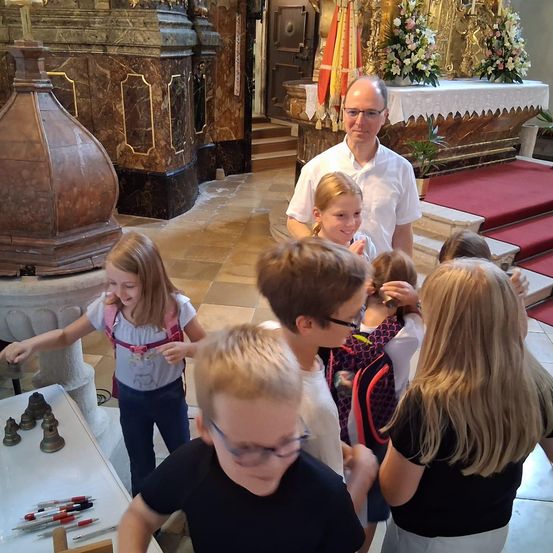 Eine Gruppe von Kindern und ein erwachsener Mann stehen in einer Kirche, in der Nähe eines Taufbeckens und eines Tisches mit Stiften und Bleistiften.