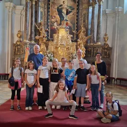 Eine Gruppe von Kindern und Erwachsenen steht vor einem kunstvollen Altar in einer Kirche und posiert für ein Foto. Der Altar hat ein großes Gemälde und goldene Statuen. Die Kinder tragen Freizeitkleidung, und einige tragen Rucksäcke.