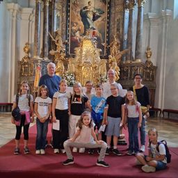 Eine Gruppe von Kindern und Erwachsenen steht vor einem kunstvollen Altar in einer Kirche und posiert für ein Foto. Der Altar hat ein großes Gemälde und goldene Statuen. Die Kinder tragen Freizeitkleidung, und einige tragen Rucksäcke.