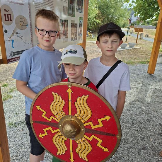 Drei junge Jungen stehen zusammen, einer hält einen roten Schild mit einem goldenen Design, während die anderen Mützen und Brillen tragen. Sie sind draußen, mit einem Plakat und einem Kiesweg hinter ihnen.