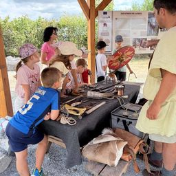 Eine Gruppe von Kindern und Erwachsenen versammelt sich um einen Tisch mit verschiedenen historischen Artefakten, möglicherweise in einer Outdoor-Bildungsumgebung. Ein Mann in antiker römischer Kleidung erklärt ihnen etwas.