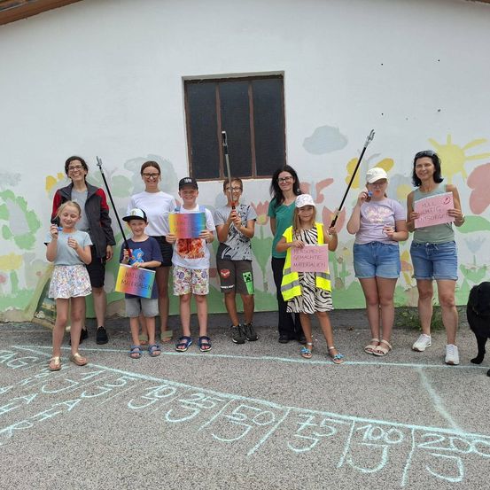 Eine Gruppe von Erwachsenen und Kindern steht vor einer Wand mit Kreidezeichnungen und hält Schilder mit Botschaften wie 'Natur' und 'Recycle Materialien'. Sie lächeln und posieren für ein Foto.