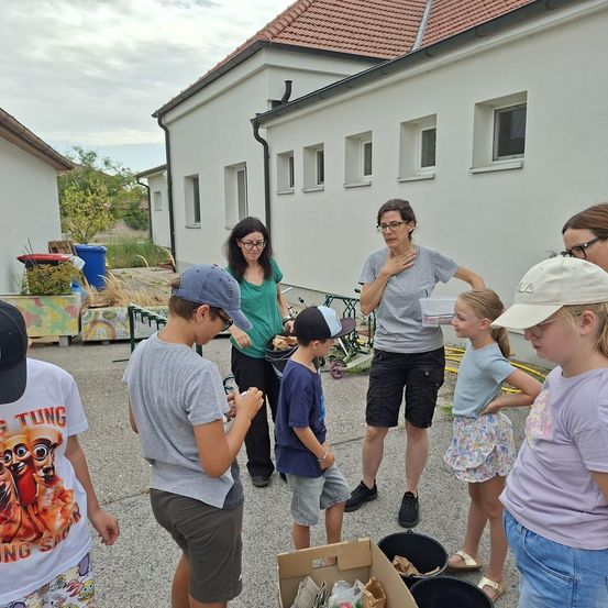 Eine Gruppe von Menschen, darunter Erwachsene und Kinder, versammelt sich vor einem Gebäude. Sie halten Essen und einige tragen Hüte. Eine Frau hält eine Schüssel und scheint mit den Kindern zu sprechen.