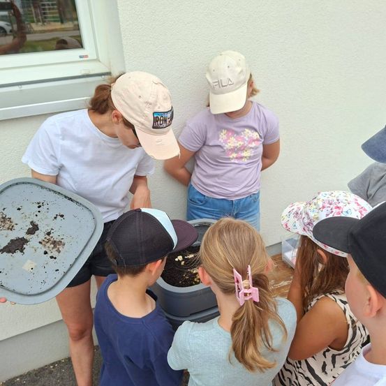Eine Gruppe von Kindern und Erwachsenen ist um einen Behälter mit Erde versammelt, möglicherweise an einer Bildungsaktivität beteiligt. Eine Frau in einem weißen T-Shirt hält eine Schale mit Erde.