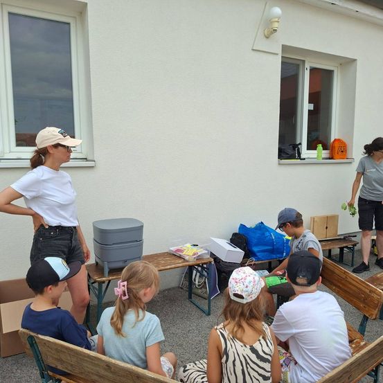 Eine Gruppe von Kindern und zwei Erwachsenen sitzen auf Bänken vor einem Gebäude. Eine Frau steht vor einem Tisch mit einem Kühler und anderen Gegenständen. Eine weitere Frau steht in der Nähe eines Fensters mit einem Rucksack und einer Flasche.
