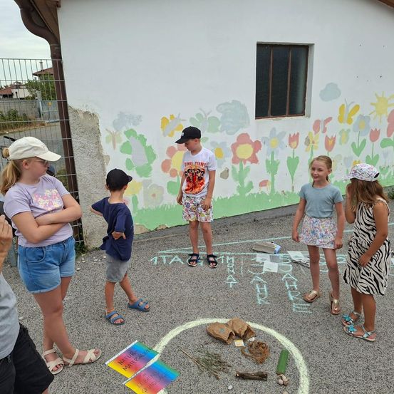 Fünf Kinder stehen in einem Kreis um eine Kreidezeichnung auf dem Boden, wobei ein Junge einen Hut und Turnschuhe trägt. Sie stehen vor einer Wand mit bunten Blumen, die darauf gemalt sind.