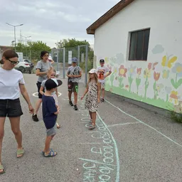 Eine Gruppe von Kindern und Erwachsenen steht in einem Kreis auf einem mit Kreide gezeichneten Pfad vor einem Gebäude mit einer Wandmalerei.