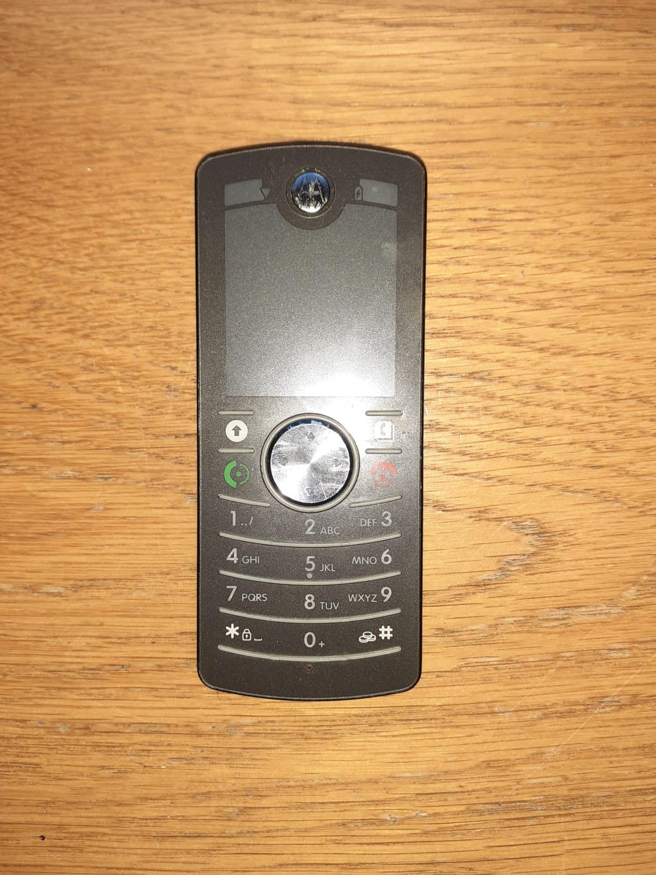 Ein Motorola-Handy mit Tastatur, auf einer Holzoberfläche platziert. Es hat eine Kamera auf der Rückseite und verschiedene Tasten für unterschiedliche Funktionen.
