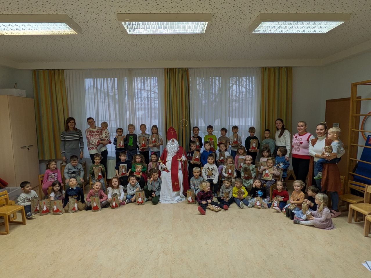 Eine Gruppe von Kindern und Erwachsenen versammelt sich in einem Raum, um ein Foto mit dem Weihnachtsmann in der Mitte zu machen, alle halten Geschenke.