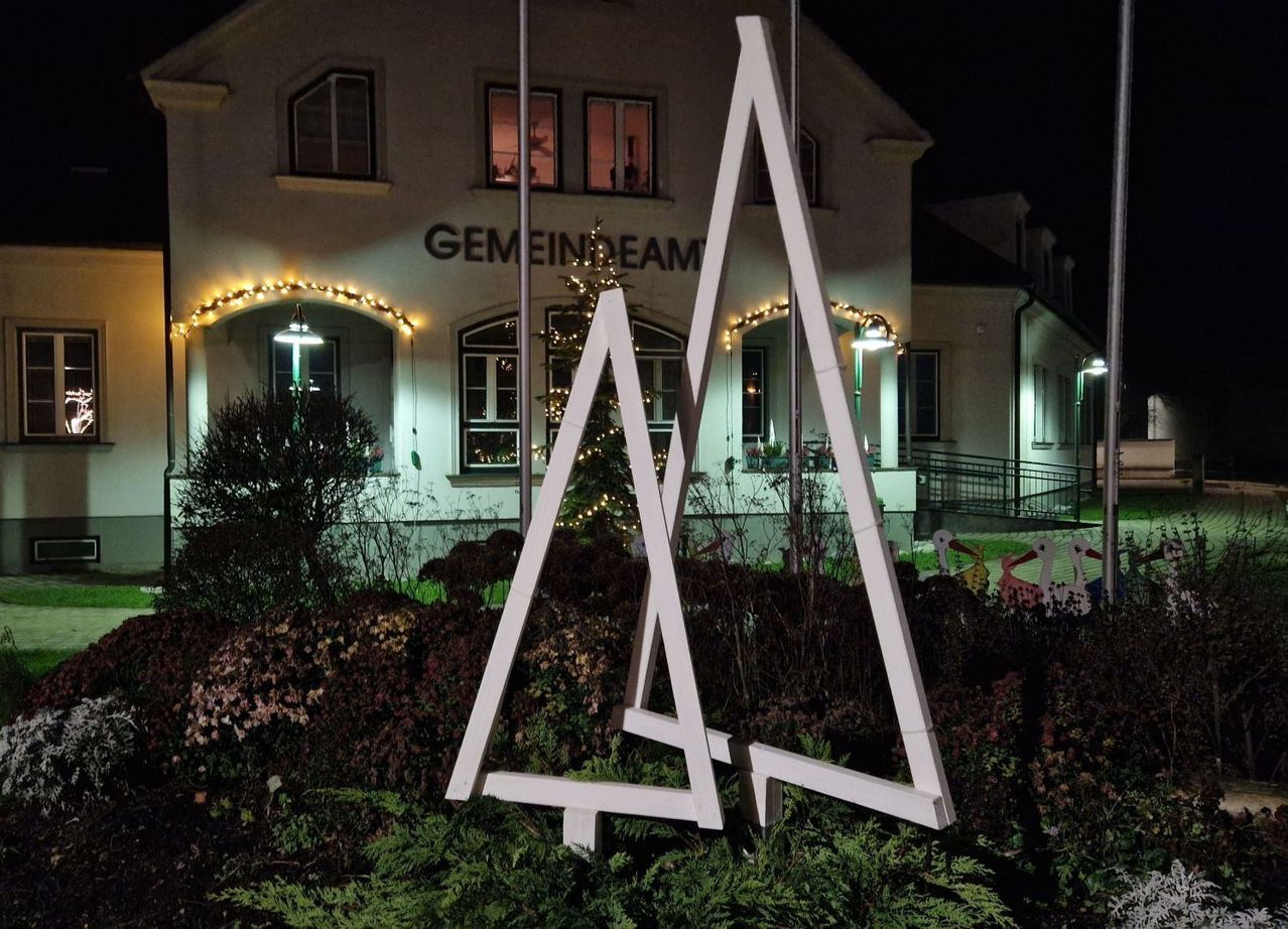 In der Nacht steht eine weiße dreieckige Struktur vor einem Gebäude mit der Aufschrift Gemeindehaus. Es gibt dekorative Lichter, einen Weihnachtsbaum und Pflanzen. Straßenlaternen stehen auf beiden Seiten des Eingangs.