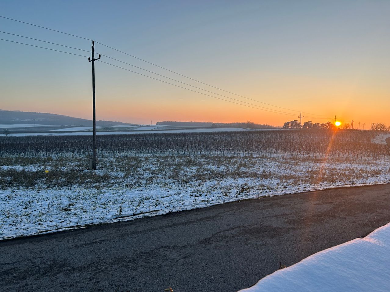 Eine verschneite Landschaft mit einer Straße, die in die Ferne führt. Stromleitungen und Masten sind sichtbar. Die Sonne geht am Horizont unter.