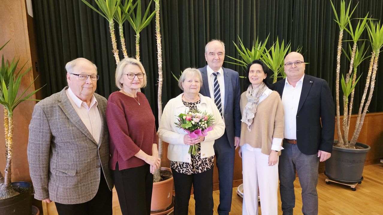 Fünf Erwachsene, darunter drei Frauen und zwei Männer, stehen zusammen in einem Raum. Die Frau in der Mitte hält einen Blumenstrauß. Sie posieren für ein Foto.