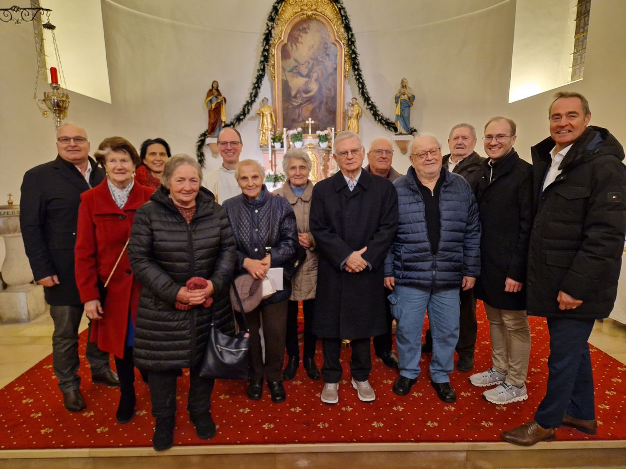 Eine Gruppe von Menschen steht vor einem Altar und lächelt für ein Foto. Dahinter schmücken Statuen und eine Girlande den Altar. Sie tragen Winterkleidung, und der Boden ist mit einem roten Teppich bedeckt.