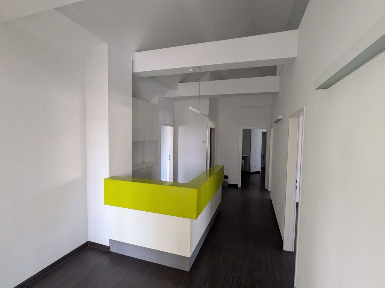 An empty modern office hallway with white walls, a yellow reception desk, and dark wooden flooring.