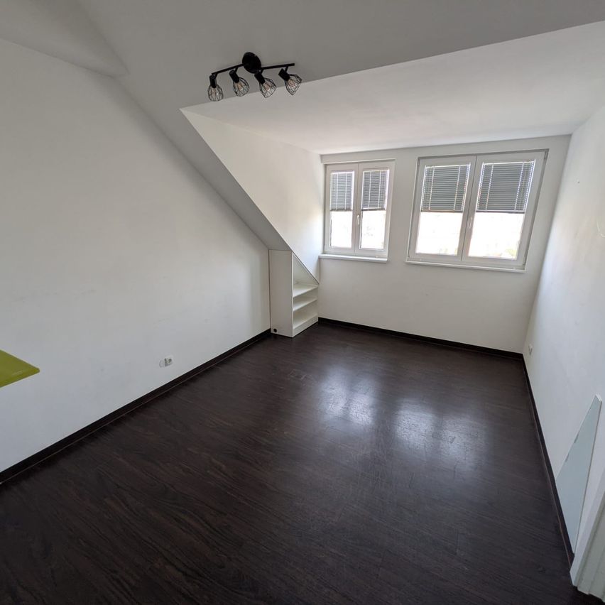 An empty attic room with white walls and dark wooden flooring. Two windows with blinds are on the right side. A small white shelf is on the left, near a wall outlet.