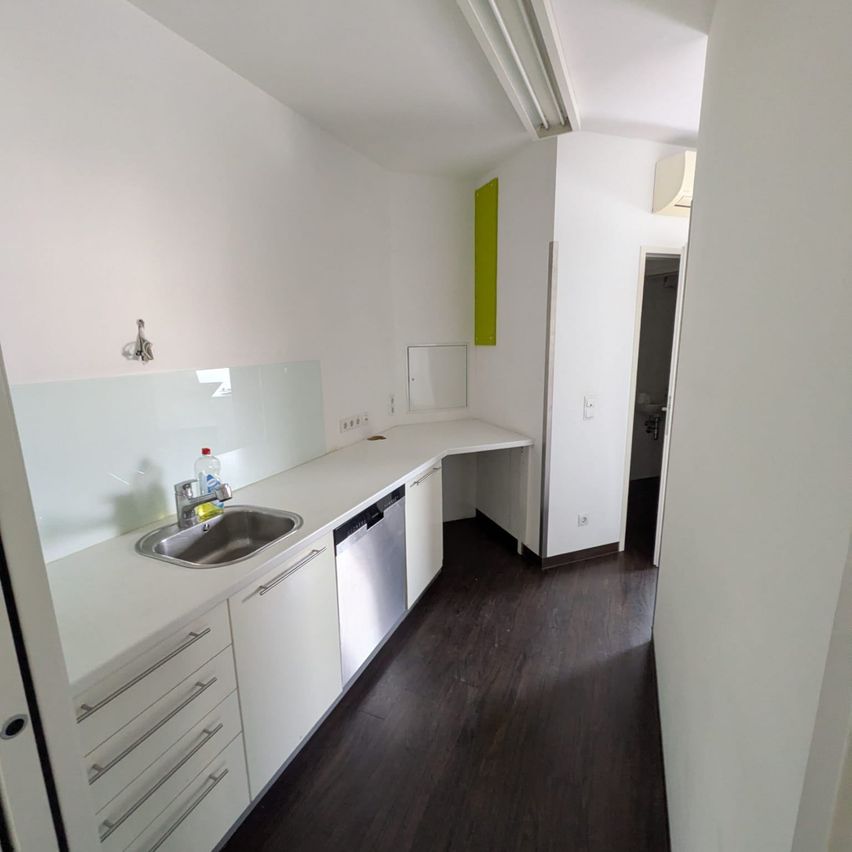 A modern kitchen with white walls and wooden floors. The countertop has a stainless steel sink with a faucet and a bottle. There are white cabinets below, and a dishwasher is beside the sink. A green wall element and a lamp are visible.