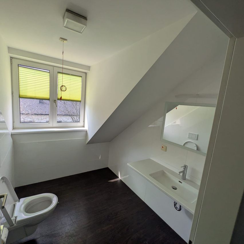 An attic bathroom with a white toilet, a sink with a faucet, and a mirror. The window has blinds and a lamp hanging from the ceiling.