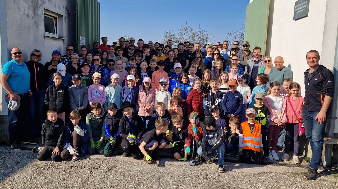 A large group of children and adults, many wearing hats and gloves, pose for a photo on a gravel surface, with trees and a building in the background.