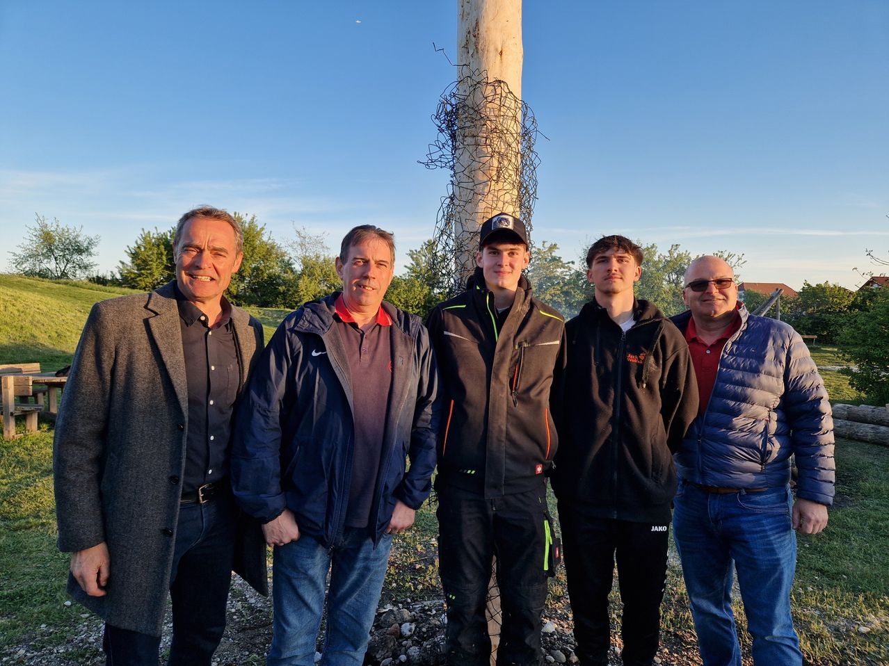 Five men stand outside near a tree, smiling for the camera. They wear jackets and jeans, with one man wearing a cap.
