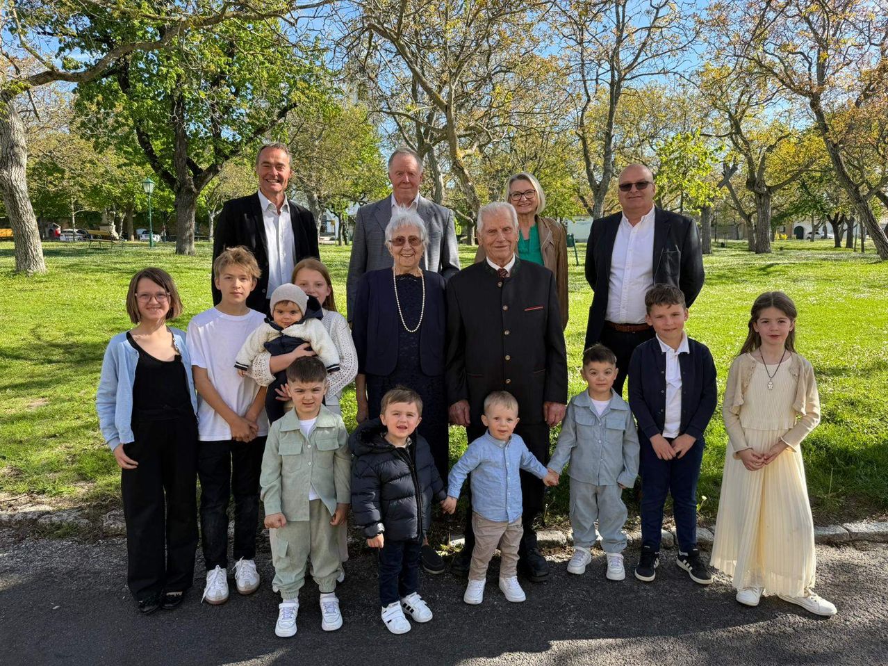 A large family poses for a photo in a park with trees in the background. Adults and children stand together on a paved road.