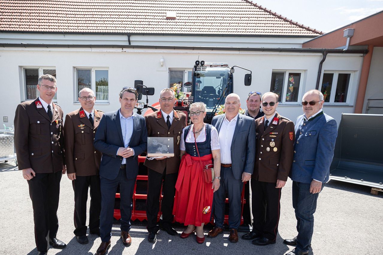 A group of people dressed in uniforms and business suits stand in front of a red tractor. One woman wears a traditional dress. They hold a plaque.