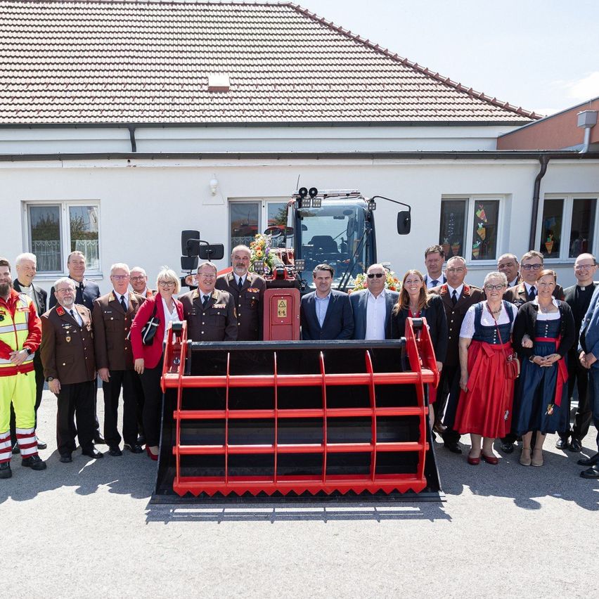 A group of people stands in front of a large red tractor. They are dressed in formal attire, and some are wearing uniforms. A building with several windows is behind them.