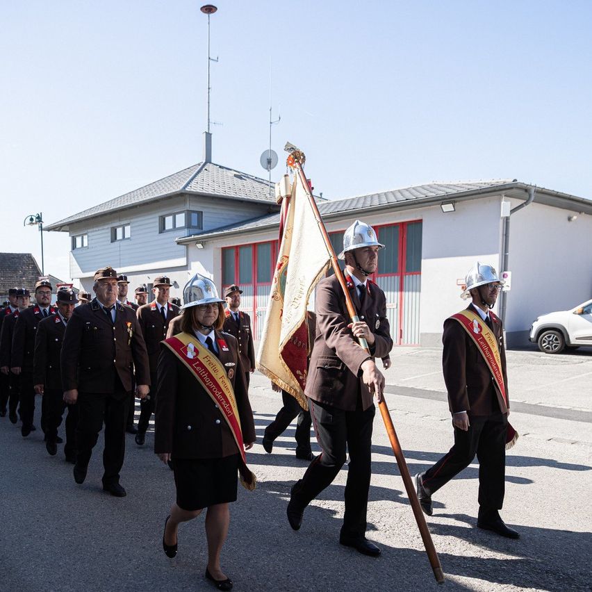 A group of firefighters in uniform, including a woman with a sash, are marching in a parade with flags and batons in front of a fire station building.