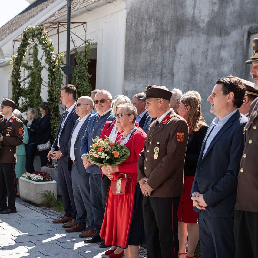 A group of people, including uniformed officials, stand in a row in front of a building, holding flowers.