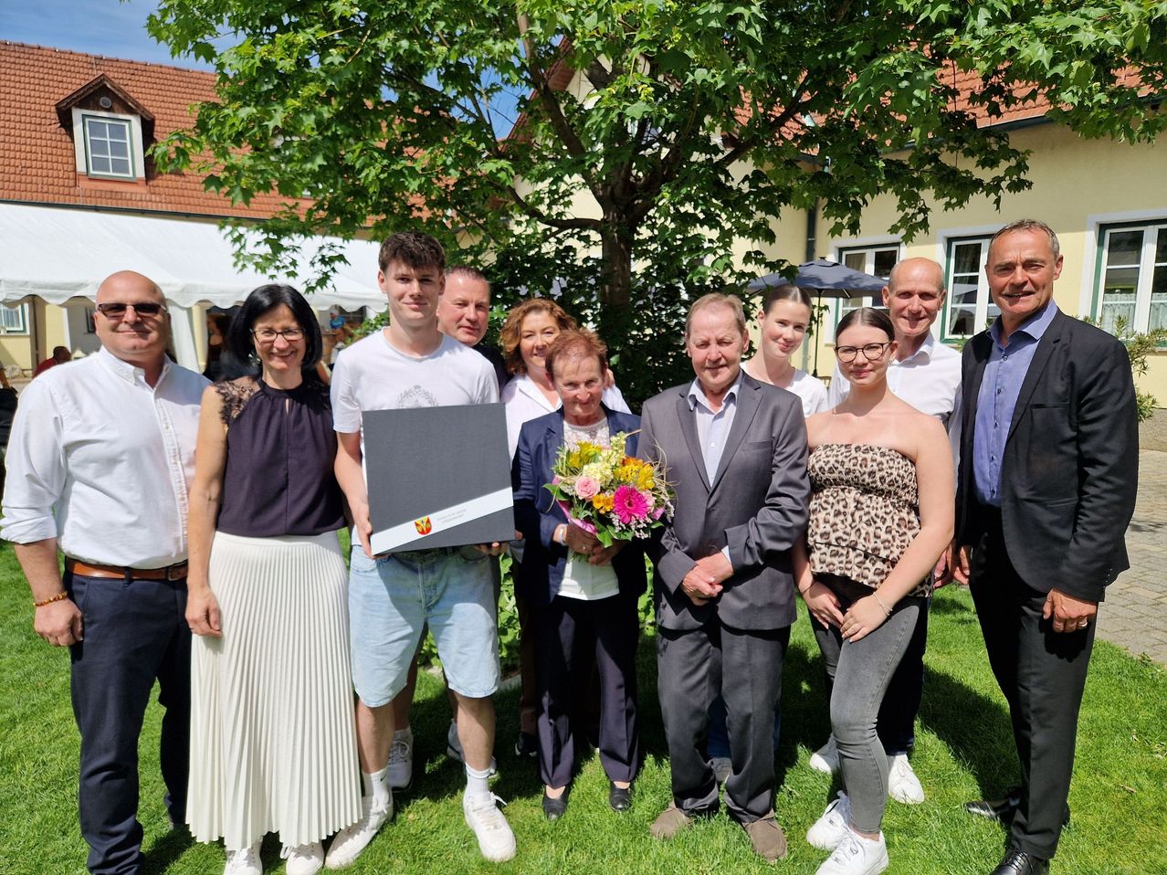Eine Gruppe von Menschen steht auf einer Wiese und posiert für ein Foto mit einem Baum im Hintergrund. Ein Mann hält ein Zertifikat und einen Blumenstrauß.