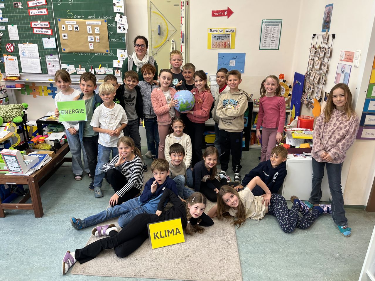 Ein Klassenfoto mit Kindern, die Schilder mit den Aufschriften Energie und Klima halten. Eine Frau steht hinter ihnen, und ein Schüler hält eine Erde. Verschiedene Klassenmaterialien und Poster schmücken die Wände.