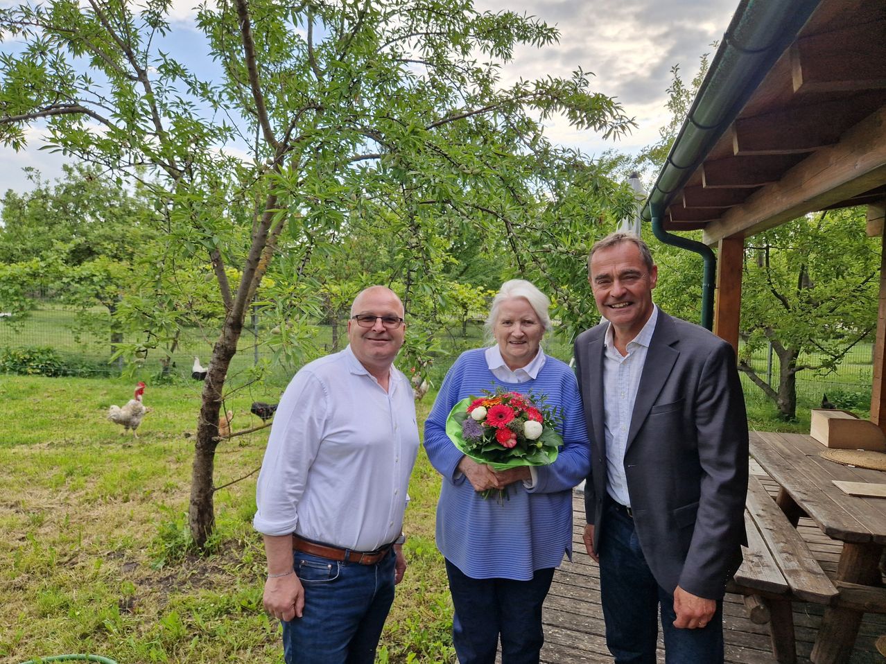 Drei Menschen stehen lächelnd vor einer Kamera im Garten. Die Frau hält einen Blumenstrauß. Hinter ihnen sind Bäume, ein Zaun und einige Hühner.