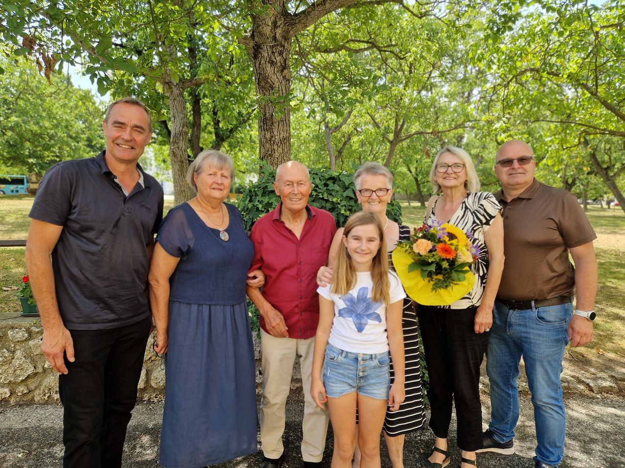 Eine Familie von sieben Personen, einschließlich eines jungen Mädchens, steht lächelnd für ein Foto in einem Park. Der Mann in der Mitte trägt ein rotes Hemd, während die Frau auf der rechten Seite einen Blumenstrauß hält.