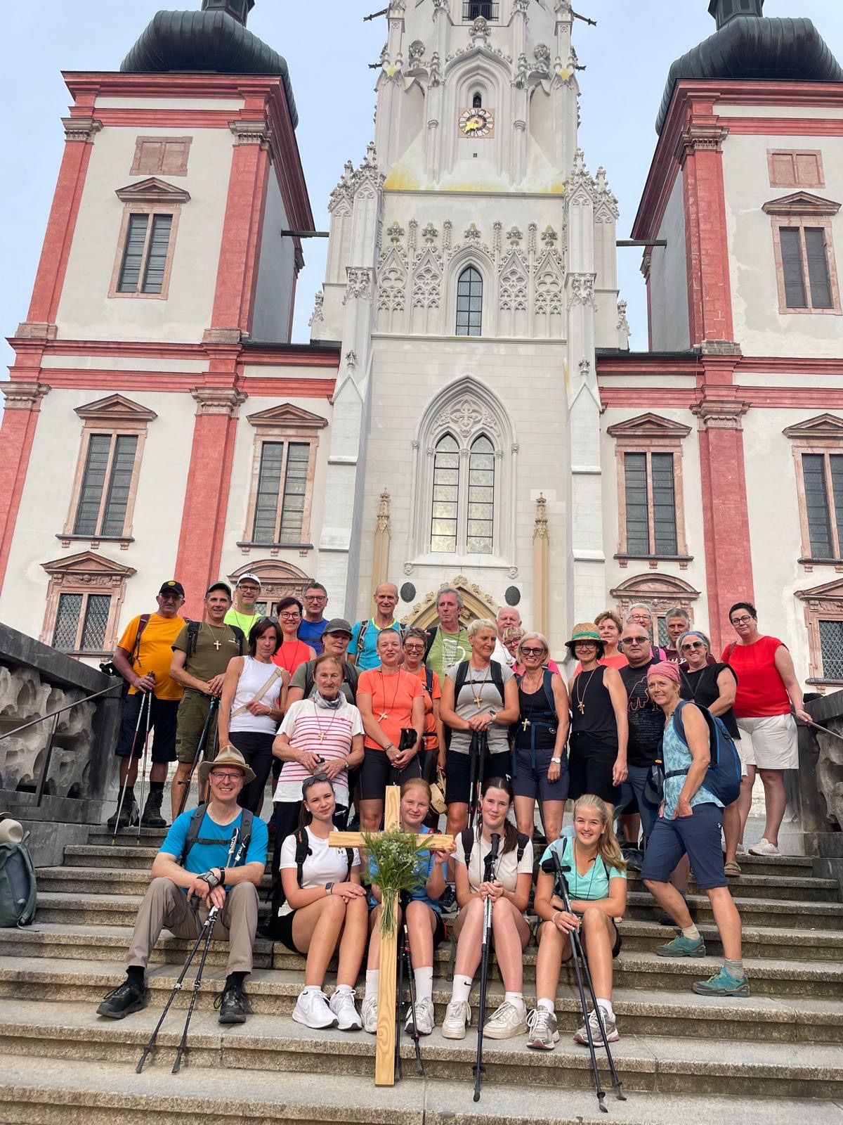 Eine Gruppe von Wanderern, einige mit Rucksäcken, posiert für ein Foto auf den Stufen einer historischen Kirche. Die Kirche weist kunstvolle Steinmetzarbeiten und einen hohen, spitzen Turm auf.
