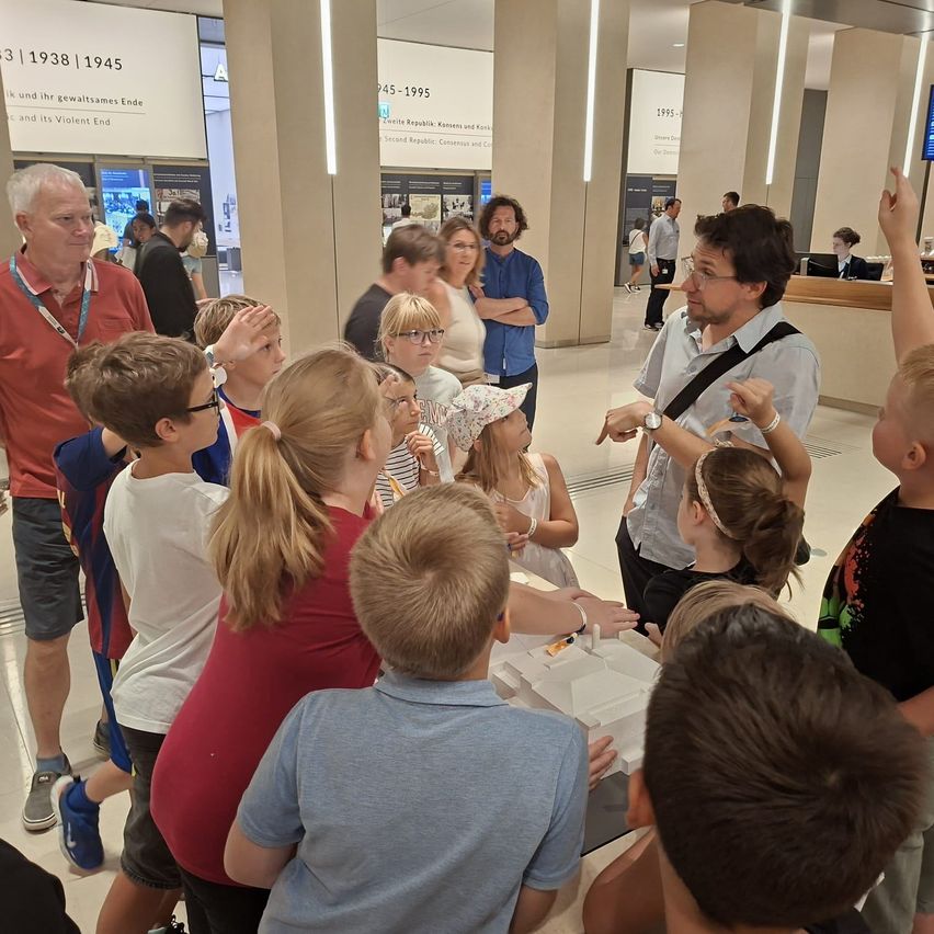 Eine Gruppe von Kindern und Erwachsenen ist um einen Mann in einem blauen Hemd versammelt, der zu ihnen spricht. Sie befinden sich in einem Museum mit Säulen und Schildern an der Wand.