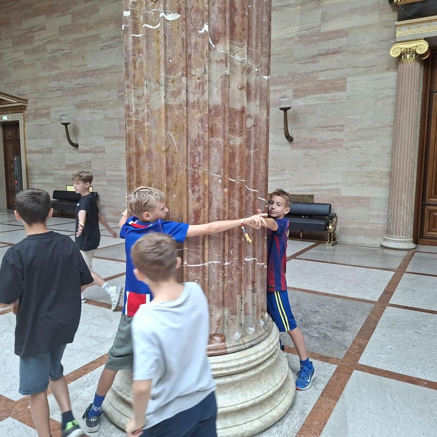 Fünf Jungen stehen um eine Marmorsäule in einem großen, prächtigen Raum. Ein Junge zeigt auf die Säule.