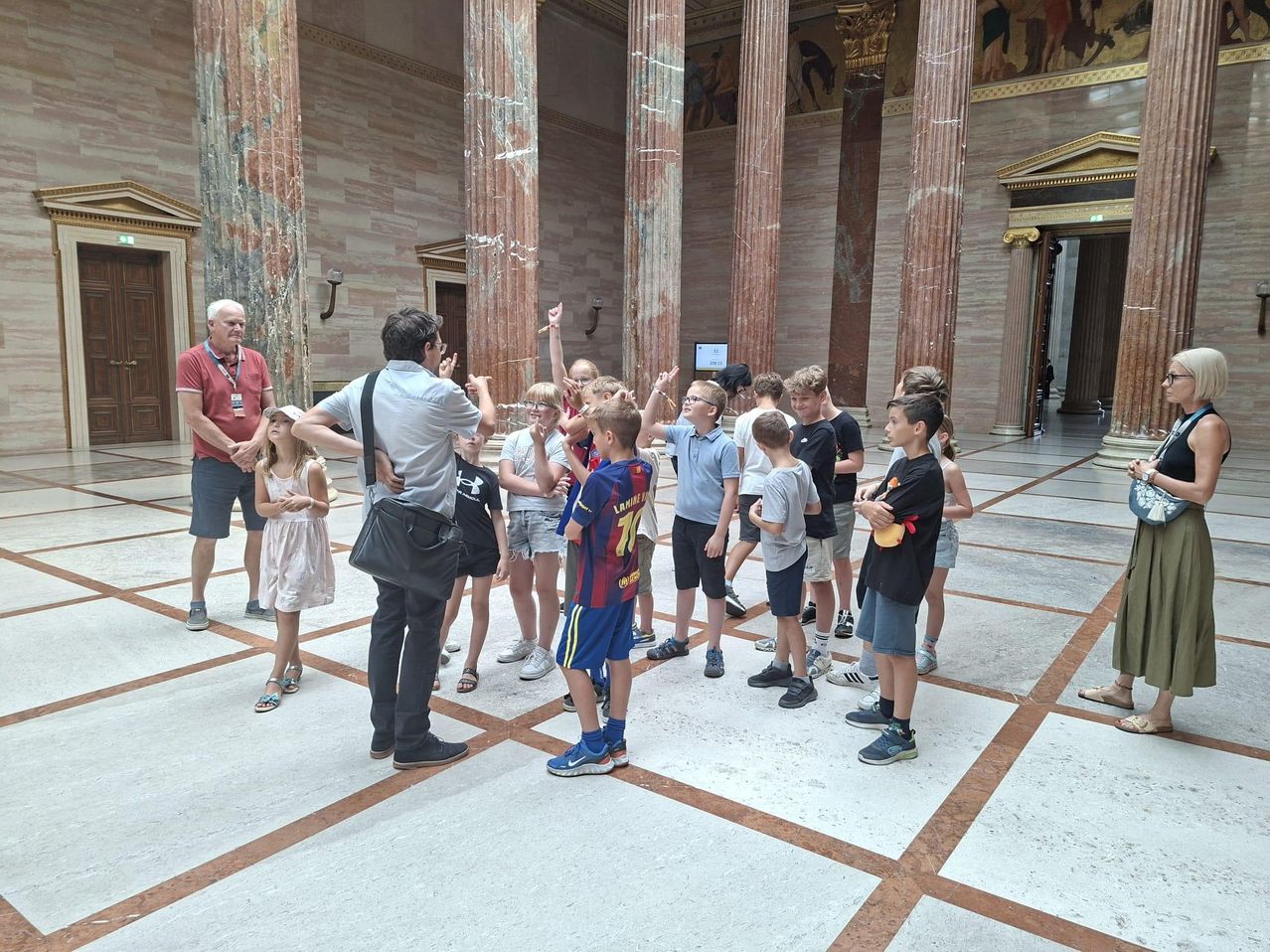 Eine Gruppe von Kindern und Erwachsenen versammelt sich in einem großen Saal mit Marmorsäulen. Die Kinder tragen Freizeitkleidung und Turnschuhe, während die Erwachsenen formeller gekleidet sind. Sie scheinen einem Führer zuzuhören.