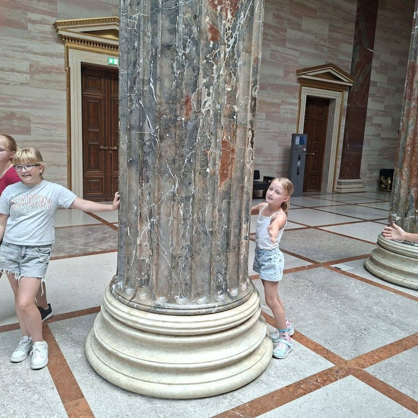 Zwei junge Mädchen stehen neben einer großen Marmorsäule in einem großen Saal, eines der Mädchen trägt eine Brille und Turnschuhe, das andere Jeansshorts und Turnschuhe.