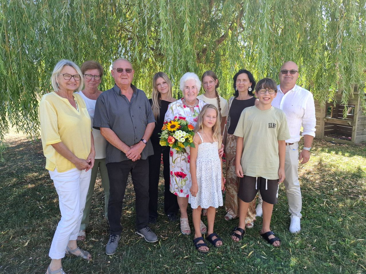 Eine Familie mit neun Personen, einschließlich Kindern, steht unter einem Weidenbaum und lächelt für ein Foto. Eine ältere Frau hält einen Blumenstrauß.