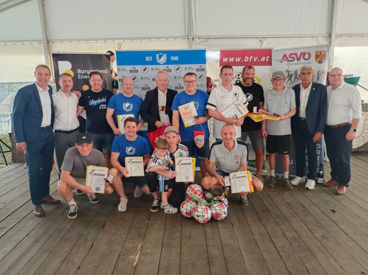 A group of men, some wearing hats and glasses, are posing for a picture with certificates and soccer balls. They stand on a wooden floor in front of a blue banner.