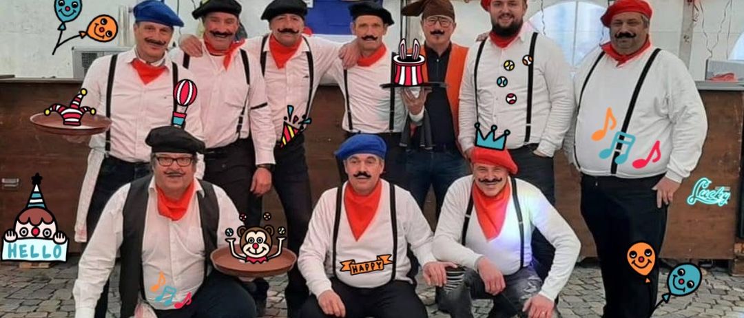 Eine Gruppe von Männern in traditioneller französischer Kleidung, die für ein Foto mit verschiedenen Requisiten posiert, darunter Hüte, Schals und ein Kuchen. Der Hintergrund zeigt eine Banner und Ballons.