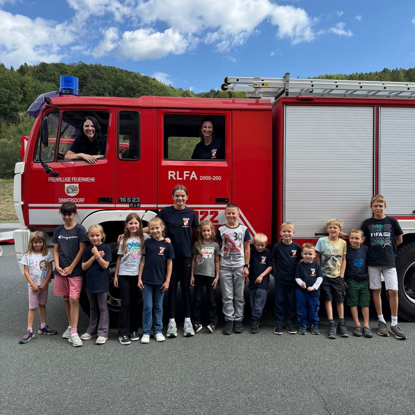 Eine Gruppe von Kindern und zwei Frauen steht vor einem roten Feuerwehrauto. Die Frauen sind im Auto und die Kinder lächeln und posieren für ein Foto.