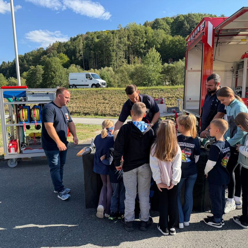 Eine Gruppe von Kindern und Erwachsenen versammelt sich um einen Feuerwehrwagen, wobei zwei Männer ihnen etwas erklären.
