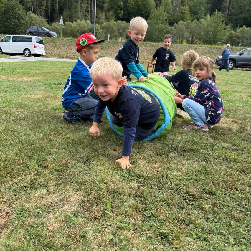 Eine Gruppe von Kindern spielt auf einem Rasenfeld, wobei eines durch eine grüne Röhre kriecht. Dahinter sind einige Autos geparkt und eine Frau steht in der Nähe eines Fahrzeugs.